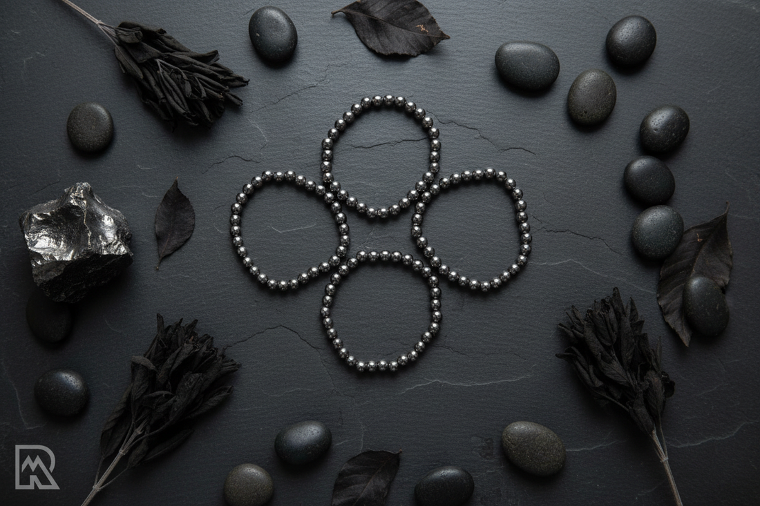 Metallic gunmetal grijze hematiet armbanden in bloempatroon op donkere leisteen met kiezelstenen, zwarte salie en ruw ijzererts in zacht dramatisch licht