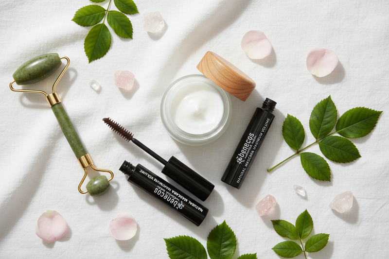 flat lay met handgemaakte gezichtscrème, een groene jade gezichtsroller en natuurlijke mascara, omringd door groene bladeren, rozenblaadjes en kleine kristallen