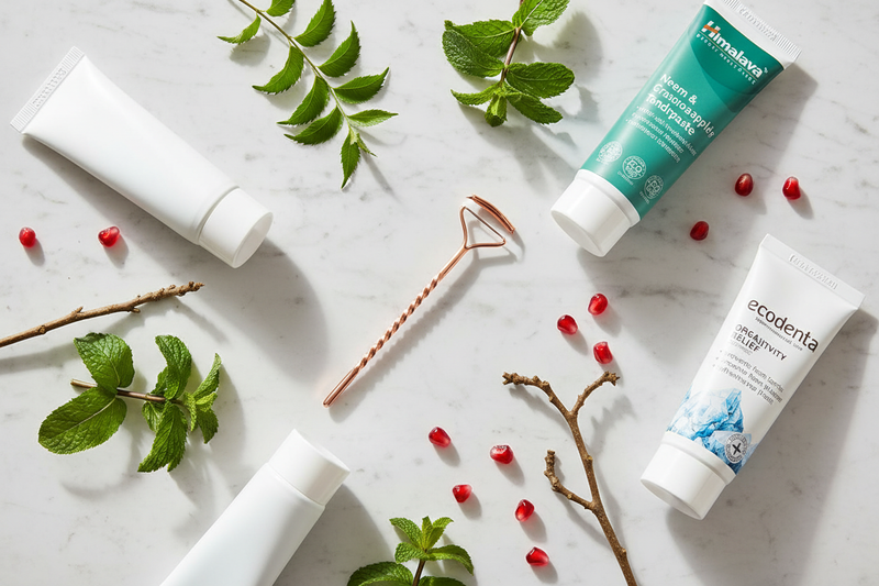 flat lay met een koperen tongschraper, natuurlijke tandpasta's en kruidenproducten op wit marmer, omringd door munt, neem en granaatappelzaadjes